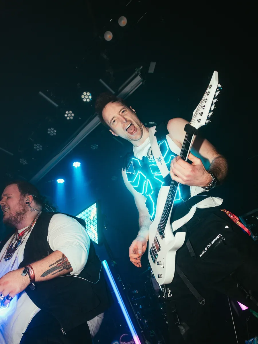 Gitarrist reisst lachend die weisse Gitarre hoch mit cyan LED-Weste